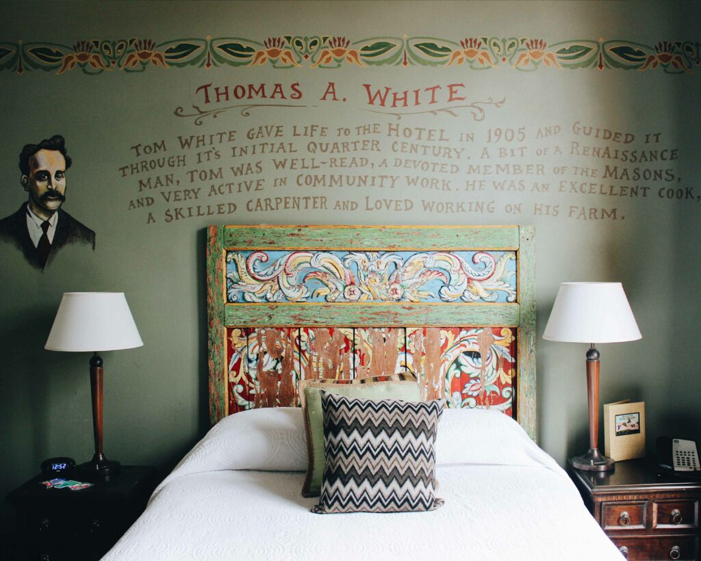 A quirky hotel bed with hand painted art on the wall behind it.  The short biography of Thomas A. White is hand-lettered on the wall.