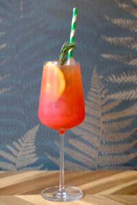 Coral colored liquid inside a wine glass. There is an orange slice, a sprig of rosemary and a green and white striped straw floating inside.