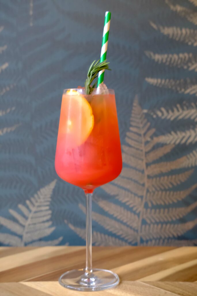 Coral colored liquid inside a wine glass.  There is an orange slice, a sprig of rosemary and a green and white striped straw floating inside.