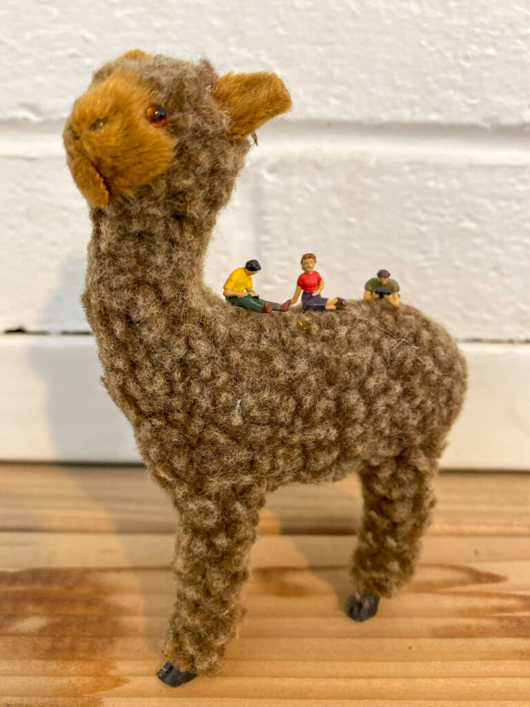 Three miniature people figurings sit atop an alpaca stuffy.