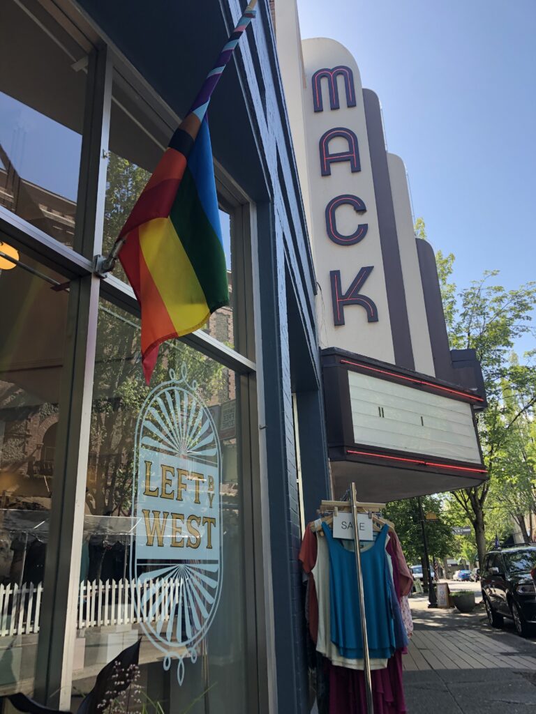 The storefront of Left By West has a pride flag displayed out front along with a sale rack.  The historic marquee of the Mack Theater is in the background.