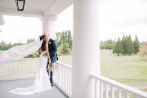 A bride and groom kiss on a large deck with white pillars.