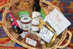 A wicker basket is full with gift items such as sourdough crackers and hummus from Collab Kitchen, Flag & Wire Coffee, Durant Olive Oil, Eola Crest Cattle Jerky, Bernards Farm Fire Roasted Hazelnuts, Free Wild She Grapefruit Bitters, Suzy's Chocolates, and Alchemist's Jam.