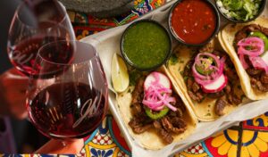 A tray of tacos and ramekins with green and red salsa. Two glasses of red wine.