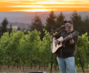 A man plays guitar and sings into a microphone. He is standing in front of a vineyard and a gorgeous sunset.