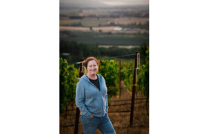 A woman in a blue hoodie stands with a vineyard behind her. She has her hands in her jean pockets and smiles.
