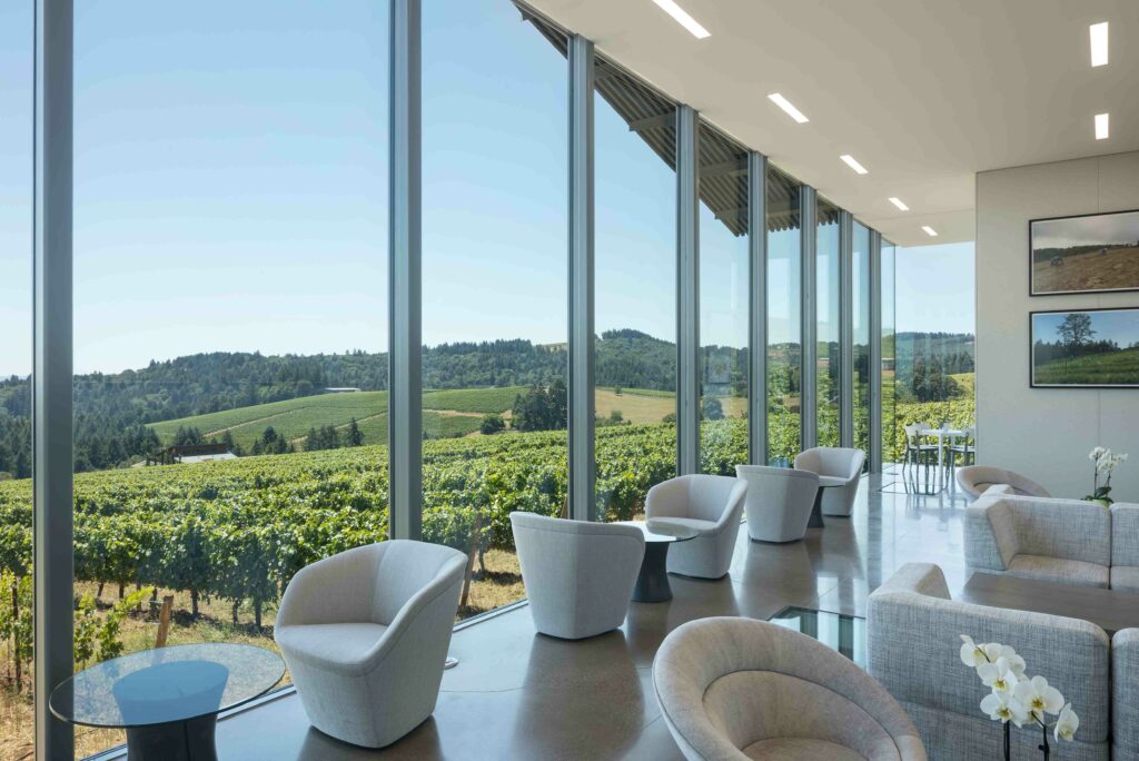 A large room with glass walls revealing sprawling vineyards. Modern white furniture and glass tables are placed throughout.