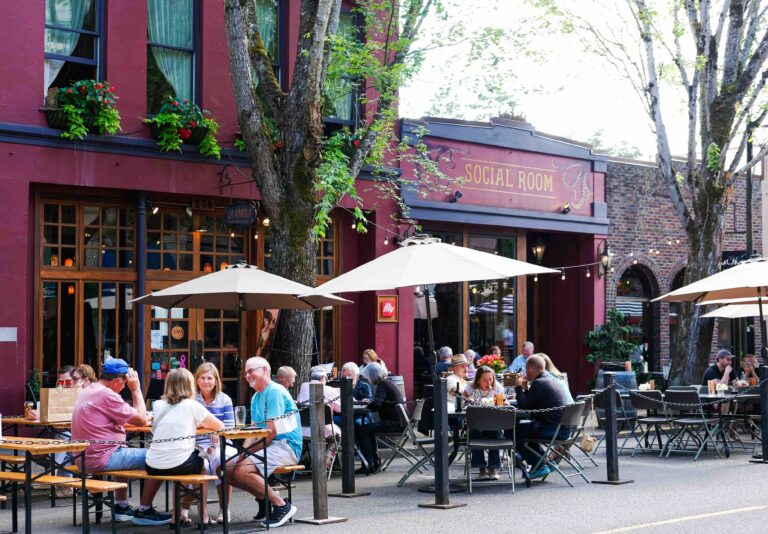 Several outdoor tables filled with people dining. They sit out in the street with the seating area roped off.