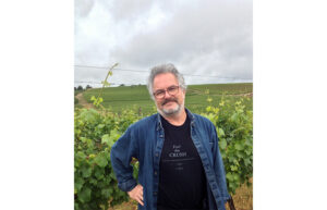 A smiling man with glasses and a beard puts his hand on his hip. He is standing in front of grape vines.