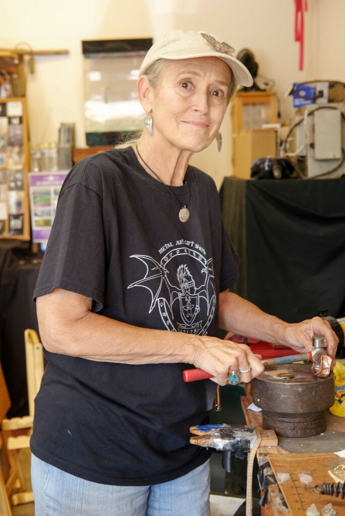 A woman with a ball cap looks at the camera and smiles.  She is holding a small hammer in one hand and a copper bracelet in the other, the bracelet resting on a small anvil.