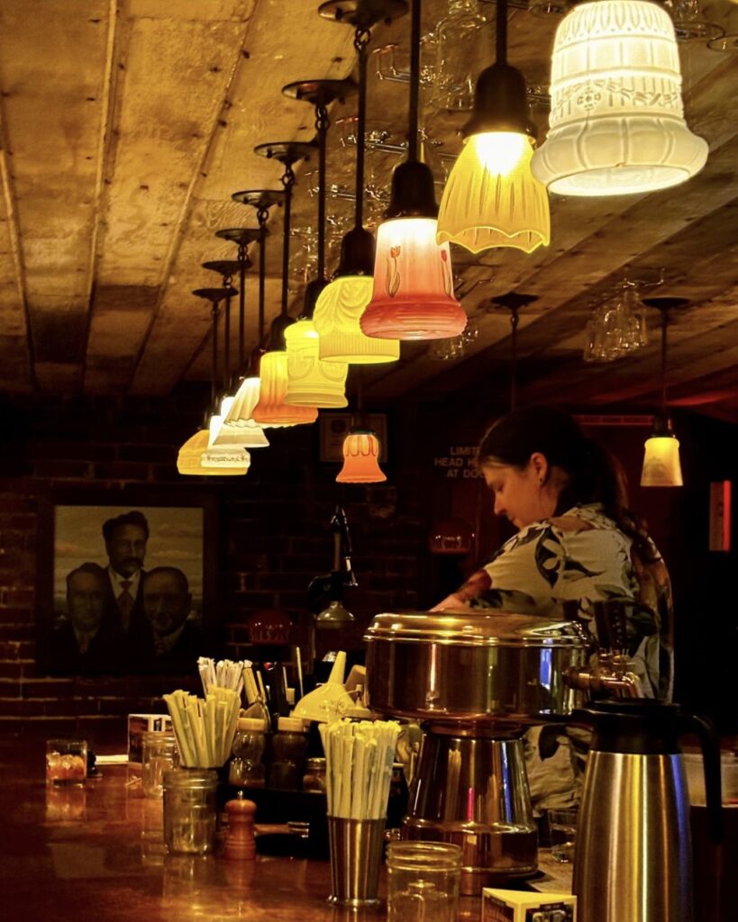 A person makes a cocktail at a warmly lit bar.  The lighting is eclectic and mismatched. The bar is made of copper.