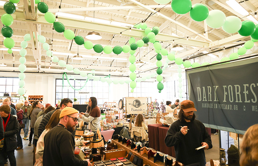 A large warehouse is well lit and painted white.  Strings of different colored green balloons are strung through the rafters.  Below, there are many tables set up with vendor's wares.  Many people are gathered, shopping.