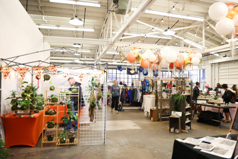 An indoor holiday market with colorful balloon displays near the ceiling and vendor booths.