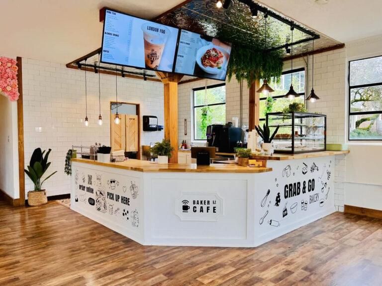 A bakery counter with a display case full of goodies. There is a tin ceiling with plants and pendant lights hanging from it. A menu is displayed on two screens.