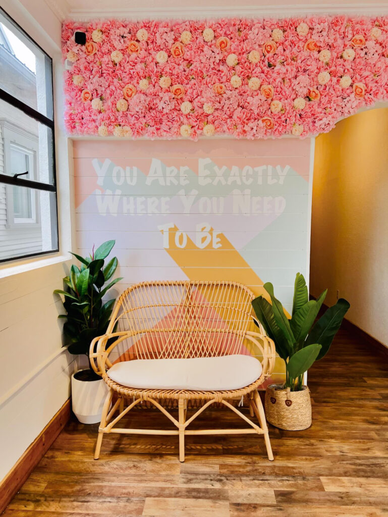 A cozy nook in the corner with a rattan bench, flanked on either side by large plants.  The wall behind it is painted to say 'You are exactly where you need to be'.  The top of the wall and archway next to the bench are covered with different shades of pink roses.
