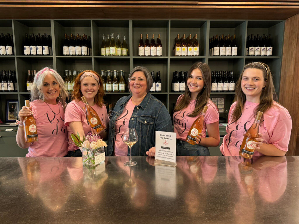 5 women with pink t-shirts that say rose all day smile and pose.  The woman in the middle has a glass of pink wine in front of her and wears a jean jacket over her pink tee.  The rest of the women hold bottles of pink Troon rose.