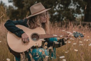 A woman sits in a field of daisies and looks at her guitar as she plays it.