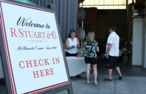 An a-frame sign says Welcome to R. Stuart & Co. Check in Here. People stand and talk at a table in the background.