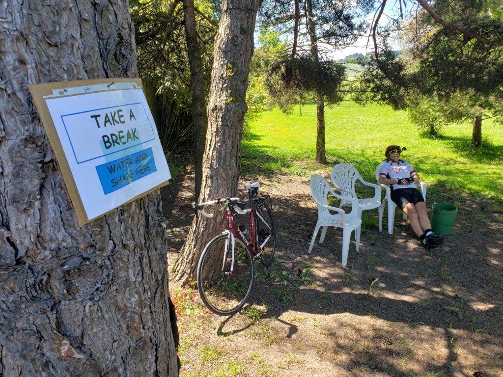 A cyclist in a helmet sits on a white outdoor chair with their bike propped up against a tree.