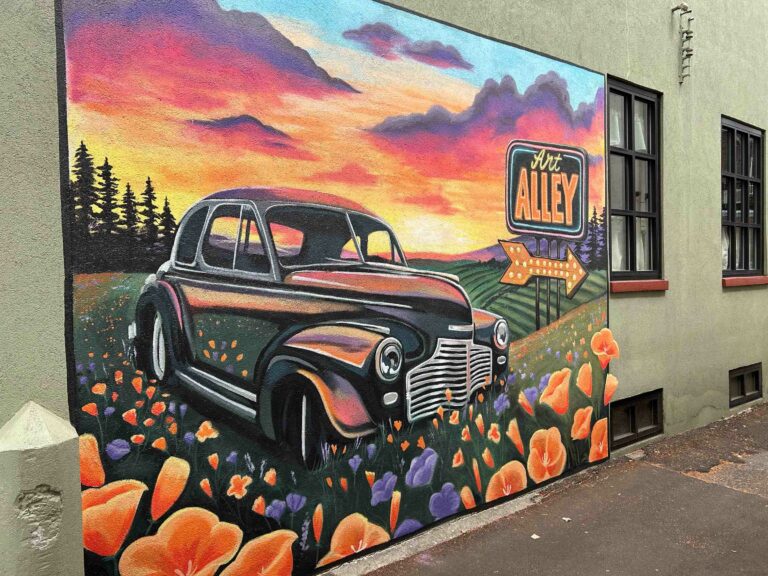 A mural of a classic car in a field of California poppies. A vibrant sunset is in the back ground along with a neon sign that says Art Alley.