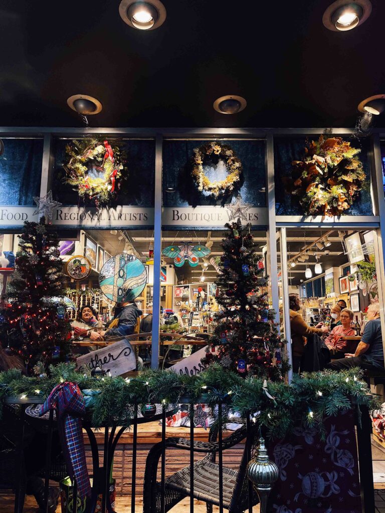 The view of a retail shop from the outside. Many people are inside, sitting at tables with bottles of wine and full glasses. There are many retail display cases behind them. The shop is warmly lit. Outside there are more tables on a patio, with Christmas trees and garland along an iron fence.