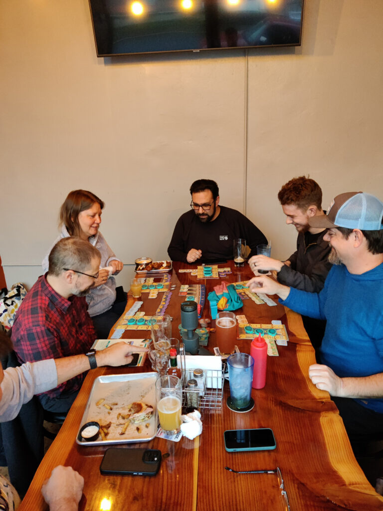 6 people focus on a board game that they are playing.  They are drinking beer and some of them have food.