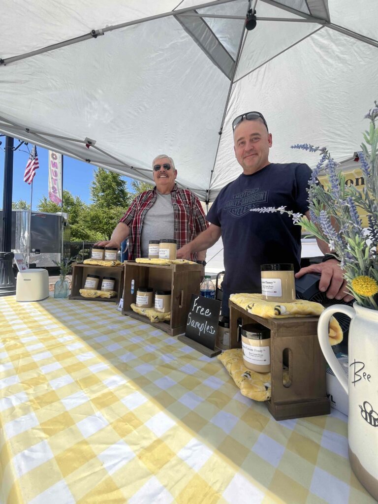Two smiling men stand underneath a tent canopy behind a table with a yellow and white table cloth.  They have displays of jars of honey and a bouquet of lavender.