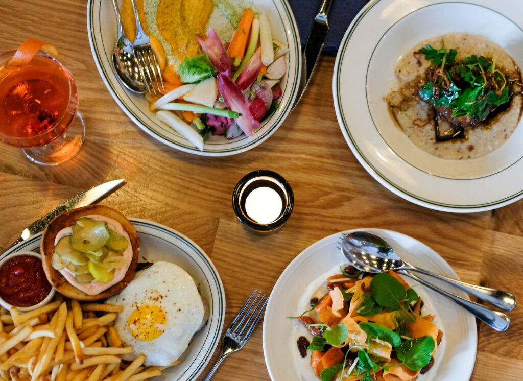 A spread of delicious looking food, including carrot hummus with fresh and pickled vegetables, a beef dish served over risotto, a hamburger with a fried egg on top and fries, and a salad with shaved carrots and pea shoots.