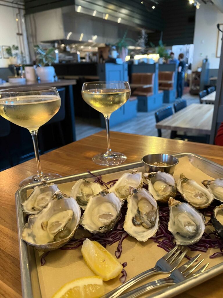Two glasses of sparkling water and a plate of fresh oyster shooters are placed on a table top.  Seating nooks are blurred in the background.