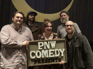A group of people smile while they hold up a sign that says PNW Comedy.