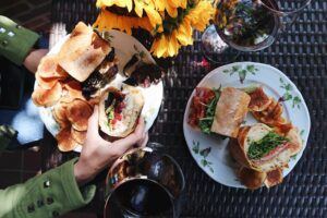 An outdoor table is set with two gourmet Italian sandwiches, chips, two glasses of wine, and a vase full of sunflowers.