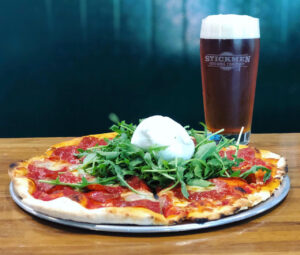 A wood fired pizza with pepperoni, arugula and a ball of burrata on top in the middle. There is a pint of beer and the glass is etched with the logo for Stickmen Brewing Company.