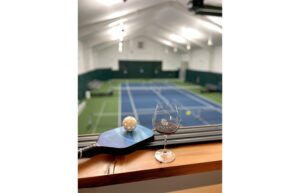 A pickleball and paddle rest next to a glass of red wine. A pickleball court is in the background.