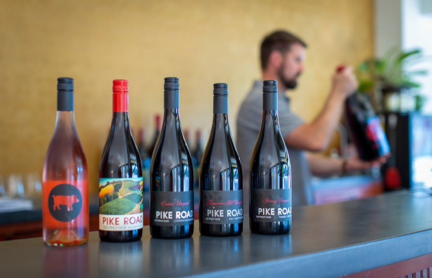 Bottles of Pike Road wine are lined up along a bar.  A blurred man holding a magnum of wine is in the background.