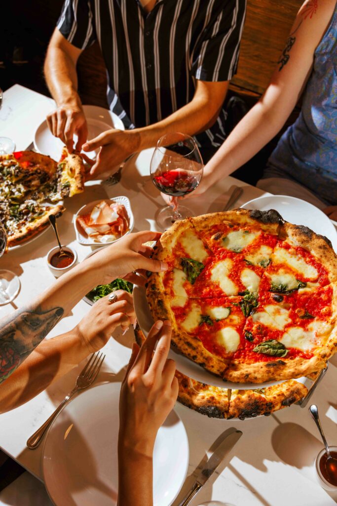 Multiples pizzas are on a table along with wine and other appetizers.  Several hands are in the photo, grabbing slices or wine glasses.