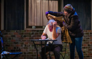 A woman in a stocking cap and coat puts a coat around a man's shoulders. They are on a stage.