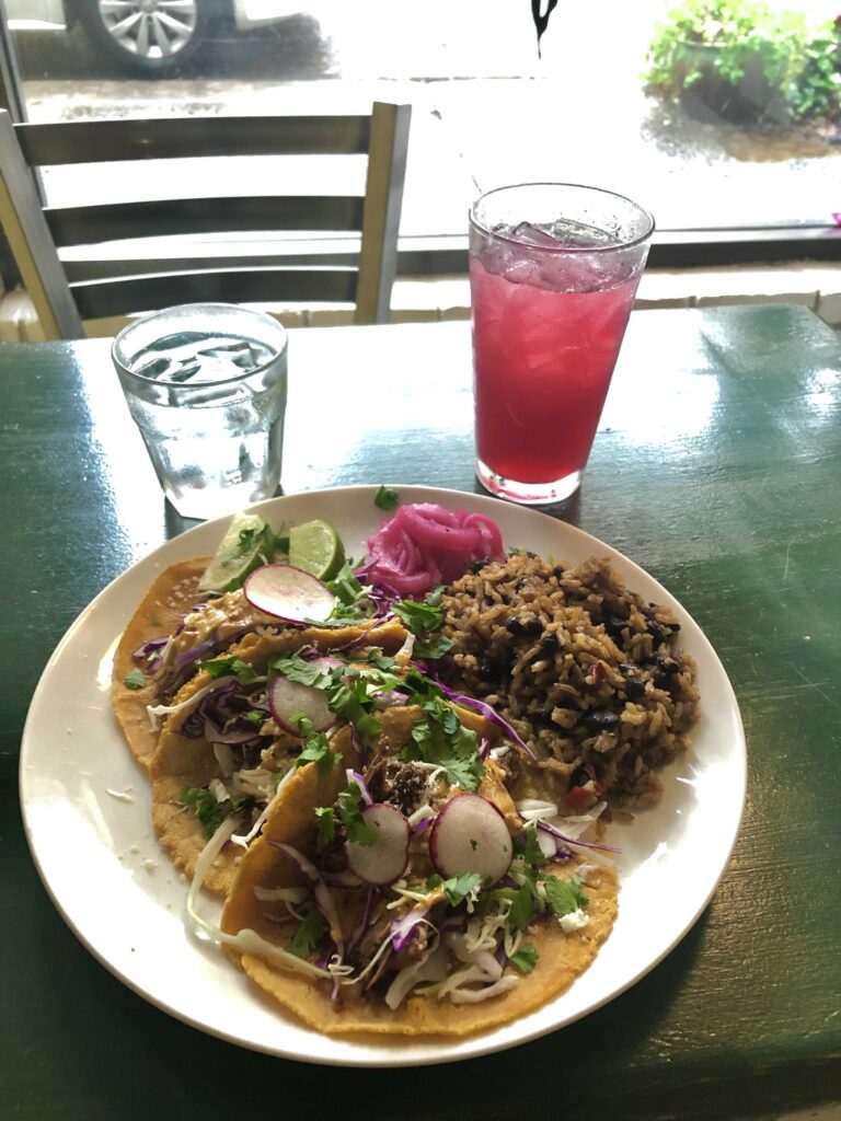 A plate with 3 tacos and rice with black beans on the side.  Purple pickled onions are the garnish. There is also a glass of something magenta to drink.