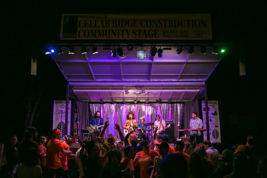 A 5 piece band plays on a lit stage at night with a lively crowd watching.