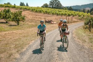 A group of cyclists ride on a gravel road with a vineyard in the background.