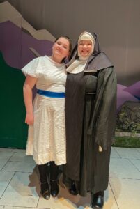 A woman smiles with her teenaged daughter. Wearing costumes, they stand in front of a set.