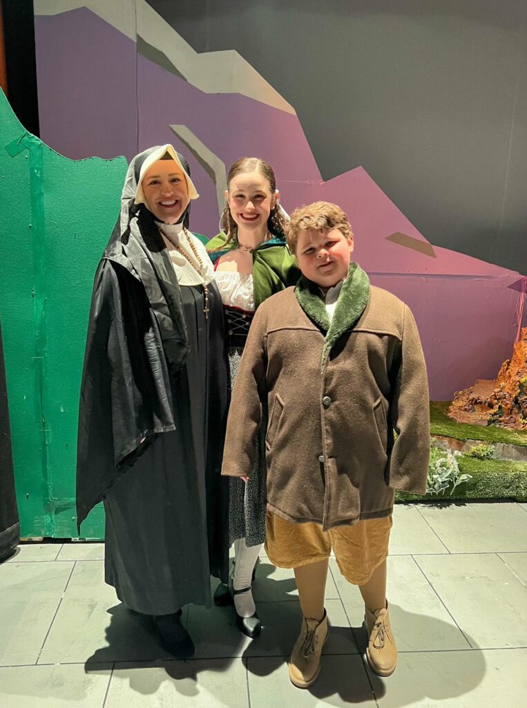 A woman smiles with her teenaged daughter and preteen son. Wearing costumes, they stand in front of a set.