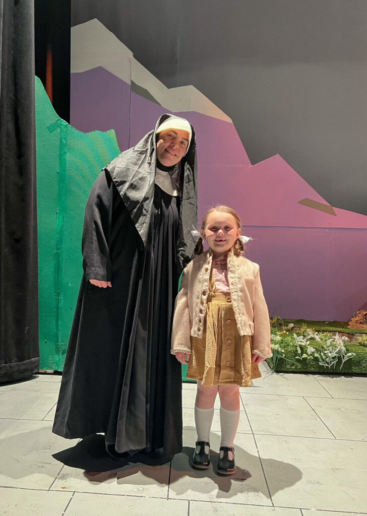 A woman smiles with her young daughter.  Wearing costumes, they stand in front of a set.