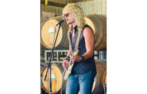 A man with long wavy hair and sunglasses plays an electric guitar and sings into a microphone. Stacked wine barrels are in the background.