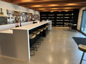 An empty bar with no back stools neatly pushed underneath. In the background there is a wall of wine.