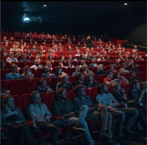 An audience in a theater sit in red seats and view a screen.