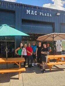 Several people stand, smiling, surrounded by picnic tables with umbrellas. Behind them is a large new looking warehouse with open garage doors. The sign on the building says Mac Plaza.