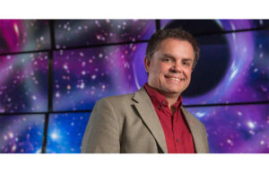 A man in a business suit smiles. He stands in front of monitors that show a colorful galaxy.