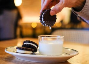 A hand holds a homemade oreo cookie dipped in milk, with a drop of milk falling down.