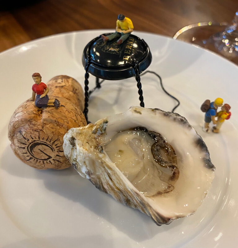 Tiny figurines sit atop a plate with an oyster, a sparkling wine cork and metal closure.