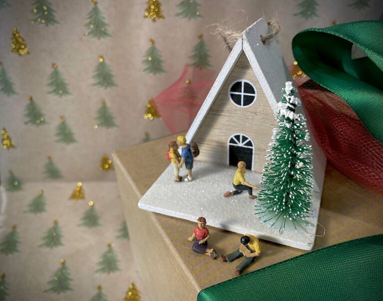 Tiny figurines are placed near a Christmas ornament of an a frame cabin with a snowy roof.